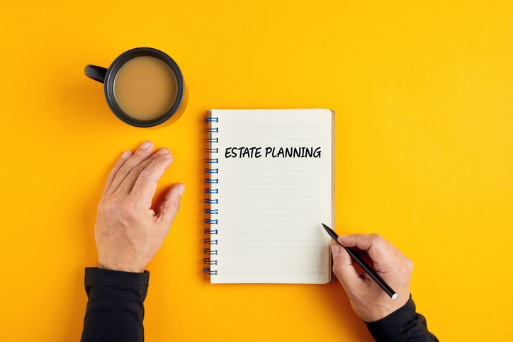 "Estate planning" written in an open notebook on top of a yellow table beside a cup of coffee.